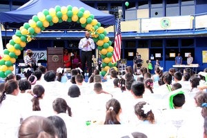A speaker addresses a crowd of children outdoors, under a balloon arch, likely at a school event or ceremony.