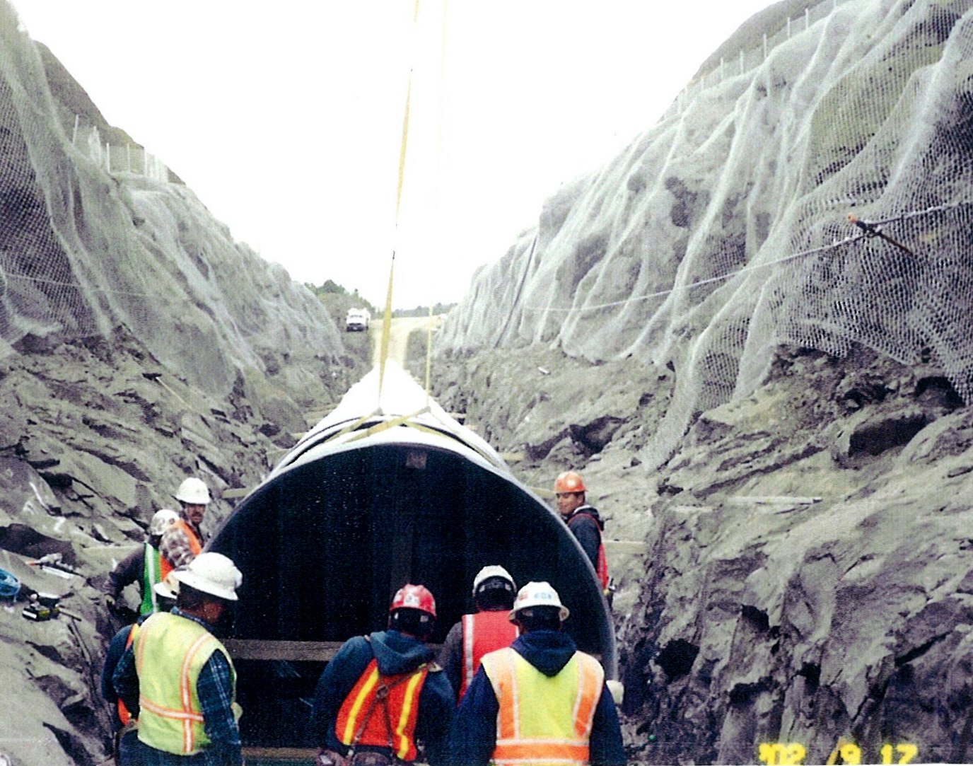 Construction workers with safety gear installing a large pipe in a rocky trench.