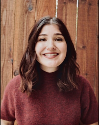 A smiling woman with brown hair wearing a burgundy sweater, standing in front of a wooden background.