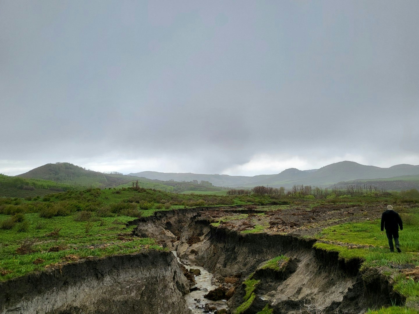 Overcast skies, hilly landscape, person near eroded land, and a small stream.