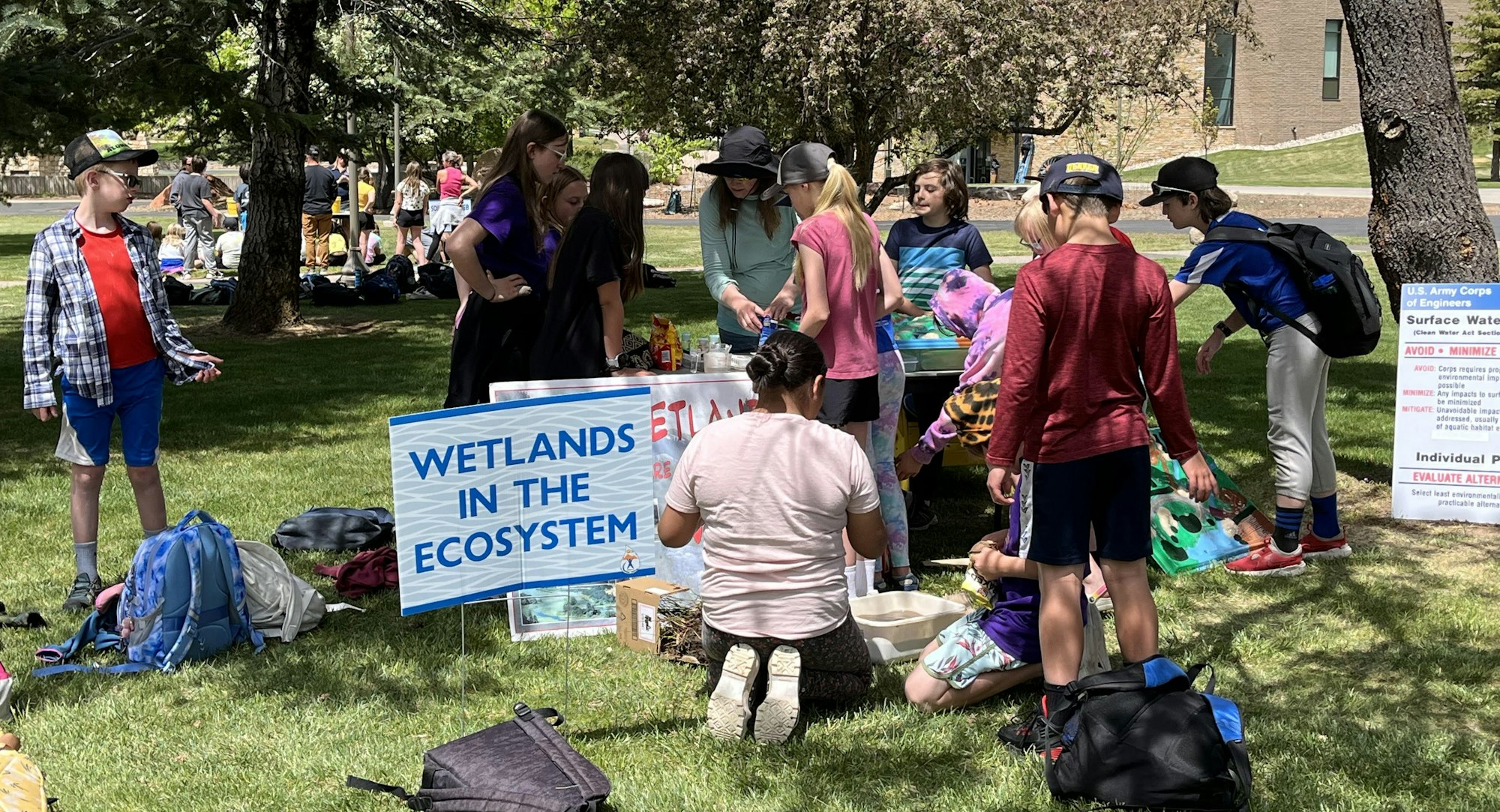 Children in a park learning about wetlands, gathered around a sign, backpacks on the grass.