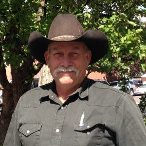 Man smiling with a mustache, wearing a cowboy hat and a dark shirt, standing in front of a tree.