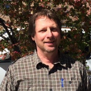 A man in a plaid shirt smiling with trees in the background.