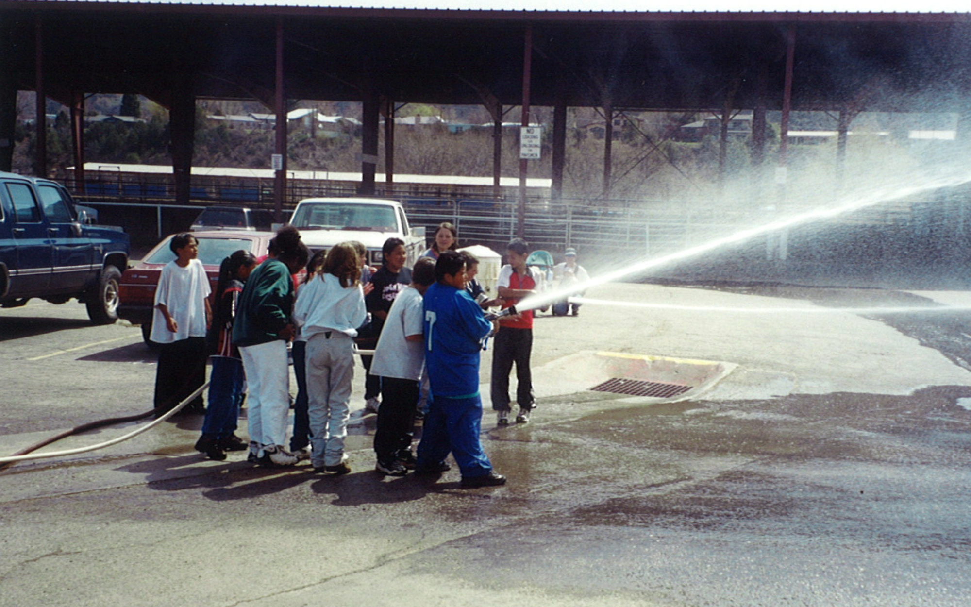Group of children watching one of them holding a fire hose, spraying water in a parking lot.