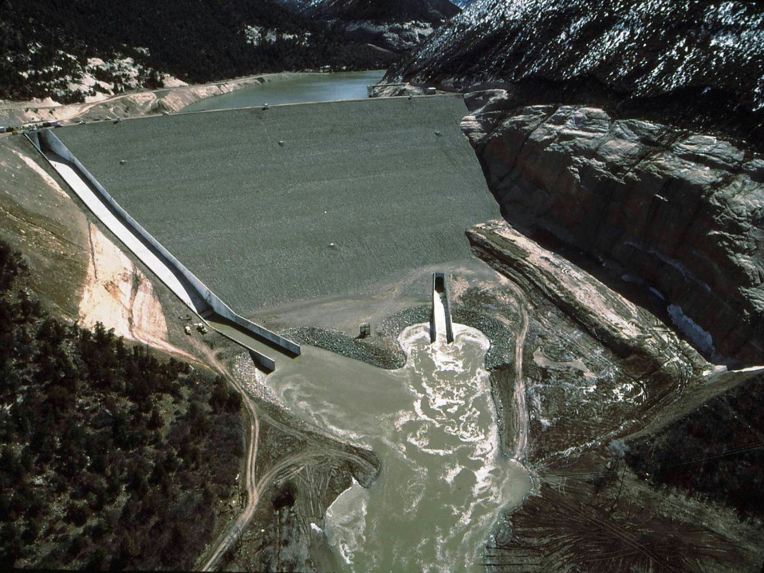 An aerial view of a dam with a reservoir behind and water discharge in front, surrounded by forested and rocky landscape.