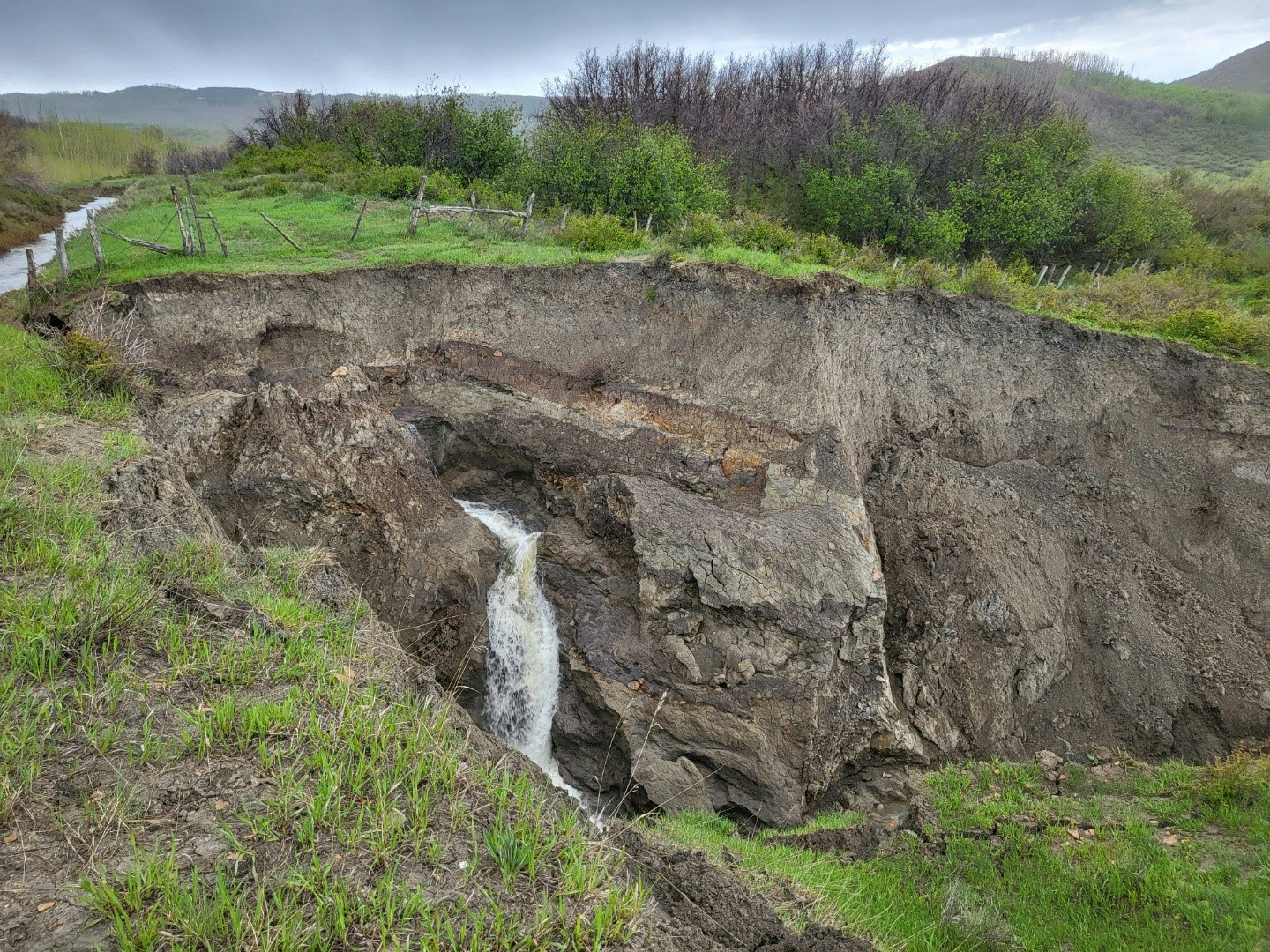 A small waterfall cascades into a rocky chasm amidst grassy, sloped terrain with distant hills and overcast skies.