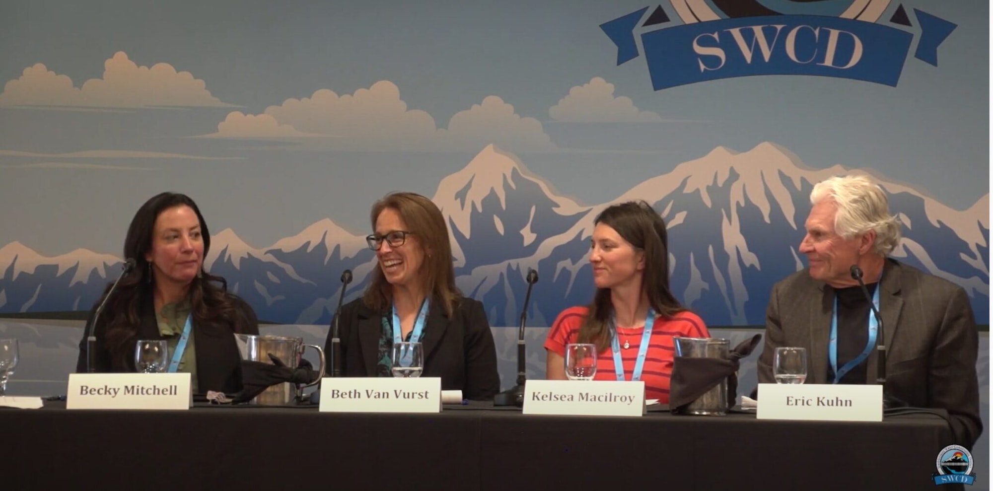 Four people at a panel discussion with name tags, sitting in front of a backdrop with mountain imagery.