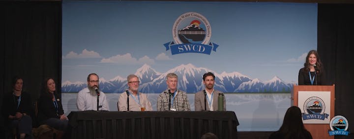 A panel of six people with one standing at a podium, in front of a backdrop with a mountain logo, at a conservation district event.
