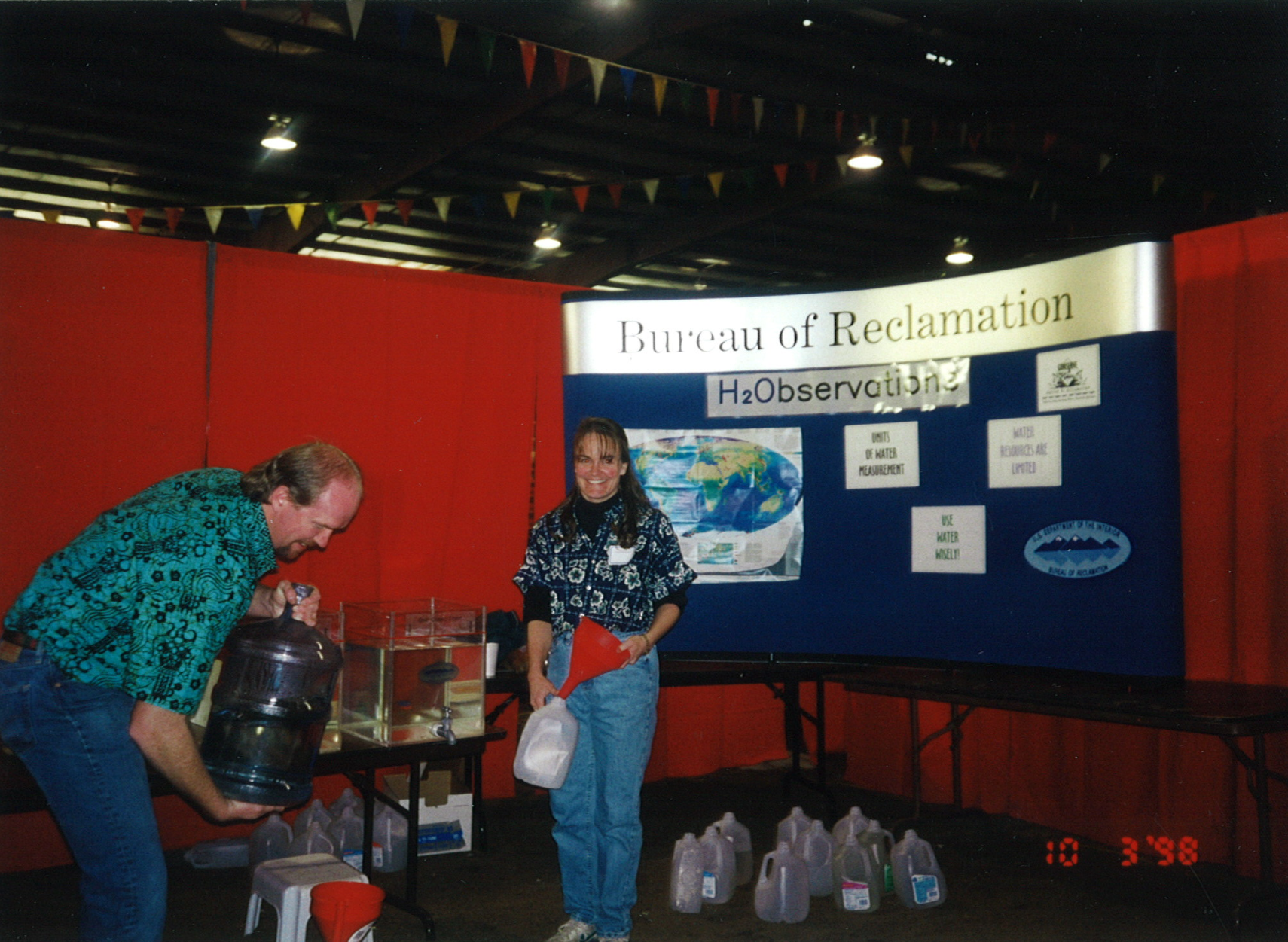 A man and a woman at a Bureau of Reclamation educational exhibit with water jugs, information, and a banner.