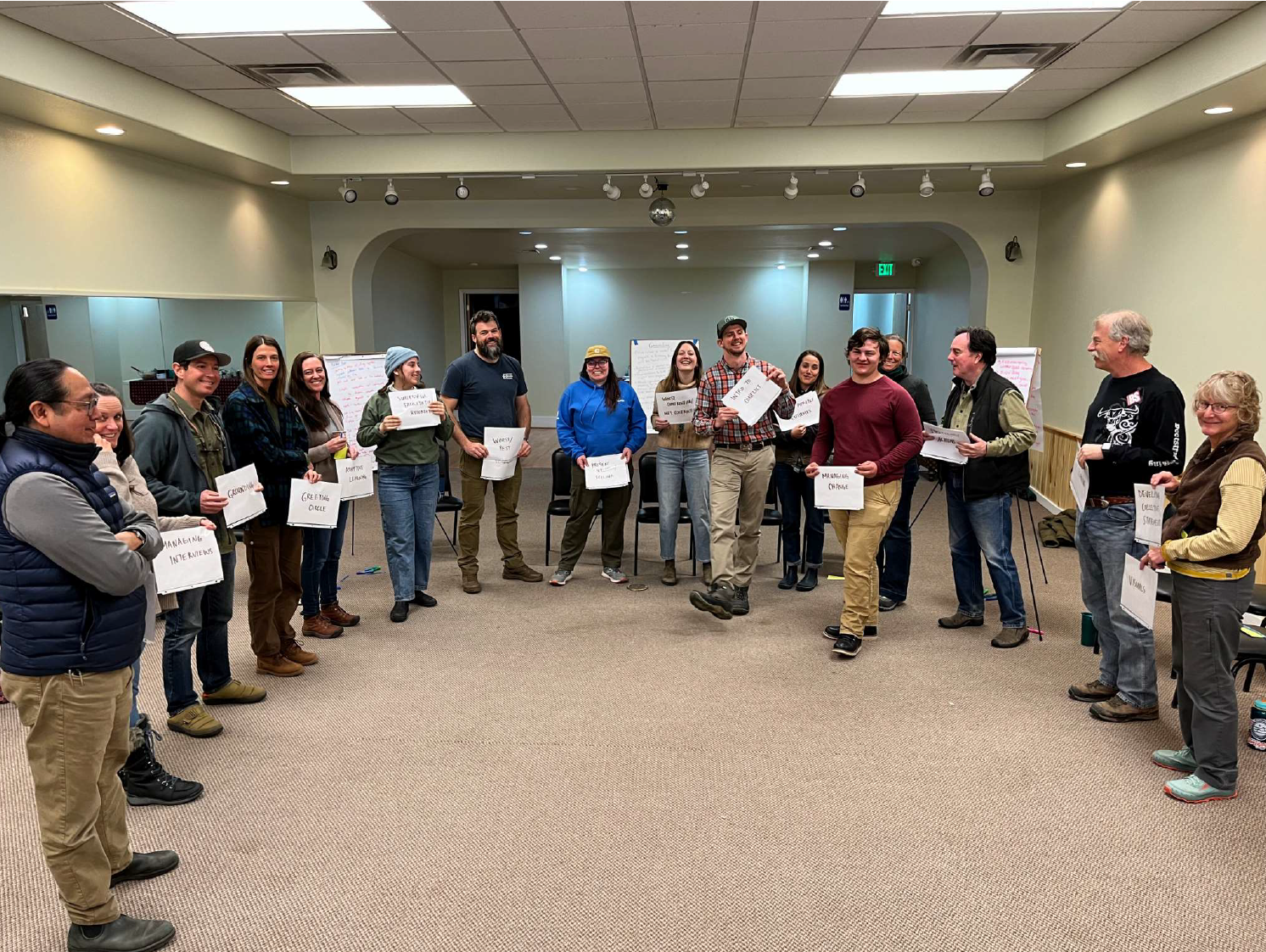 A group of people standing in a semi-circle, each holding a piece of paper, possibly in a team-building or workshop setting.