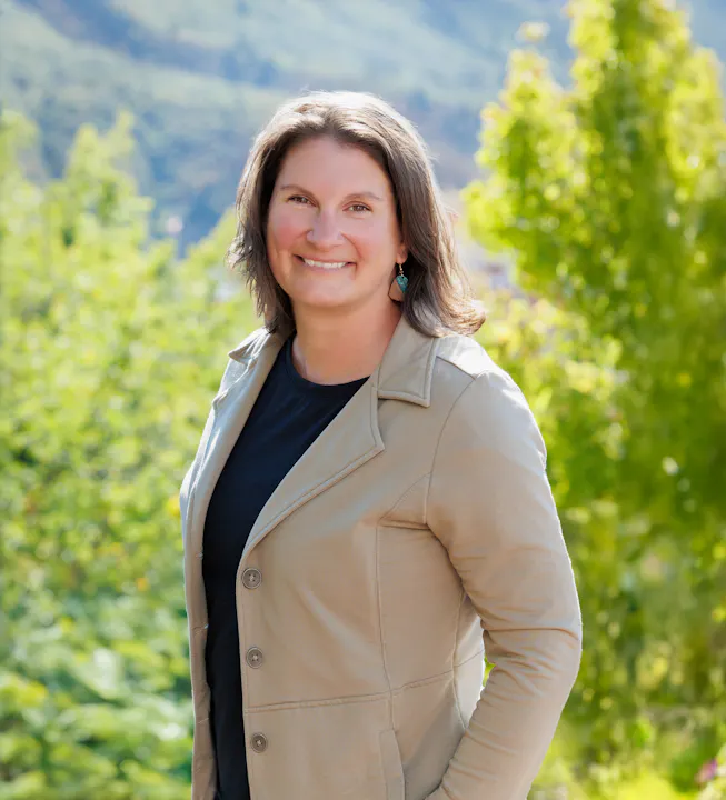 A smiling woman in a light jacket stands outdoors, surrounded by greenery and mountains in the background.