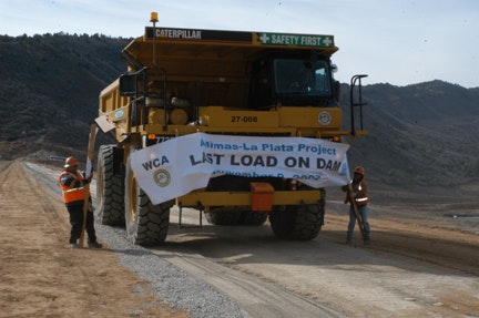 A large dump truck with a banner celebrating the "LAST LOAD ON DAM" at the Animas-La Plata Project, flanked by two workers in safety gear.