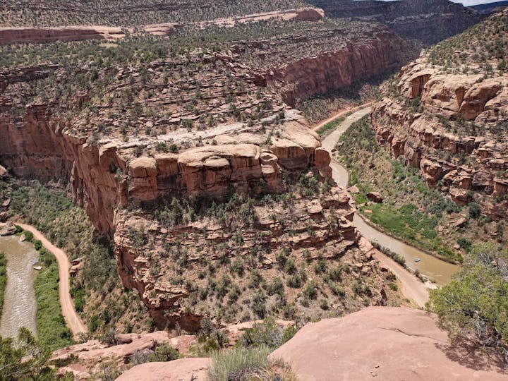 A winding river flows through a canyon with steep cliffs and green vegetation below. A dirt road runs parallel to the river.
