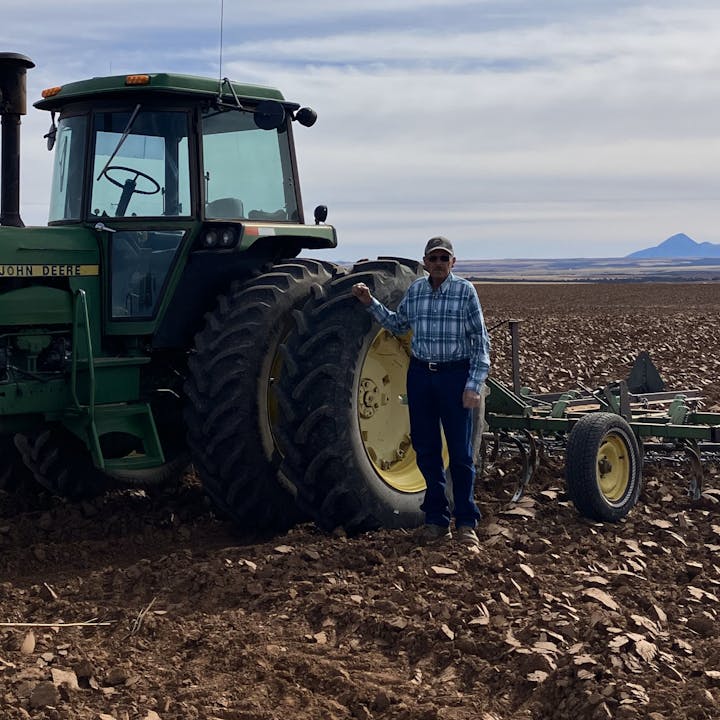 A person stands next to a John Deere tractor in a plowed field under a cloudy sky.