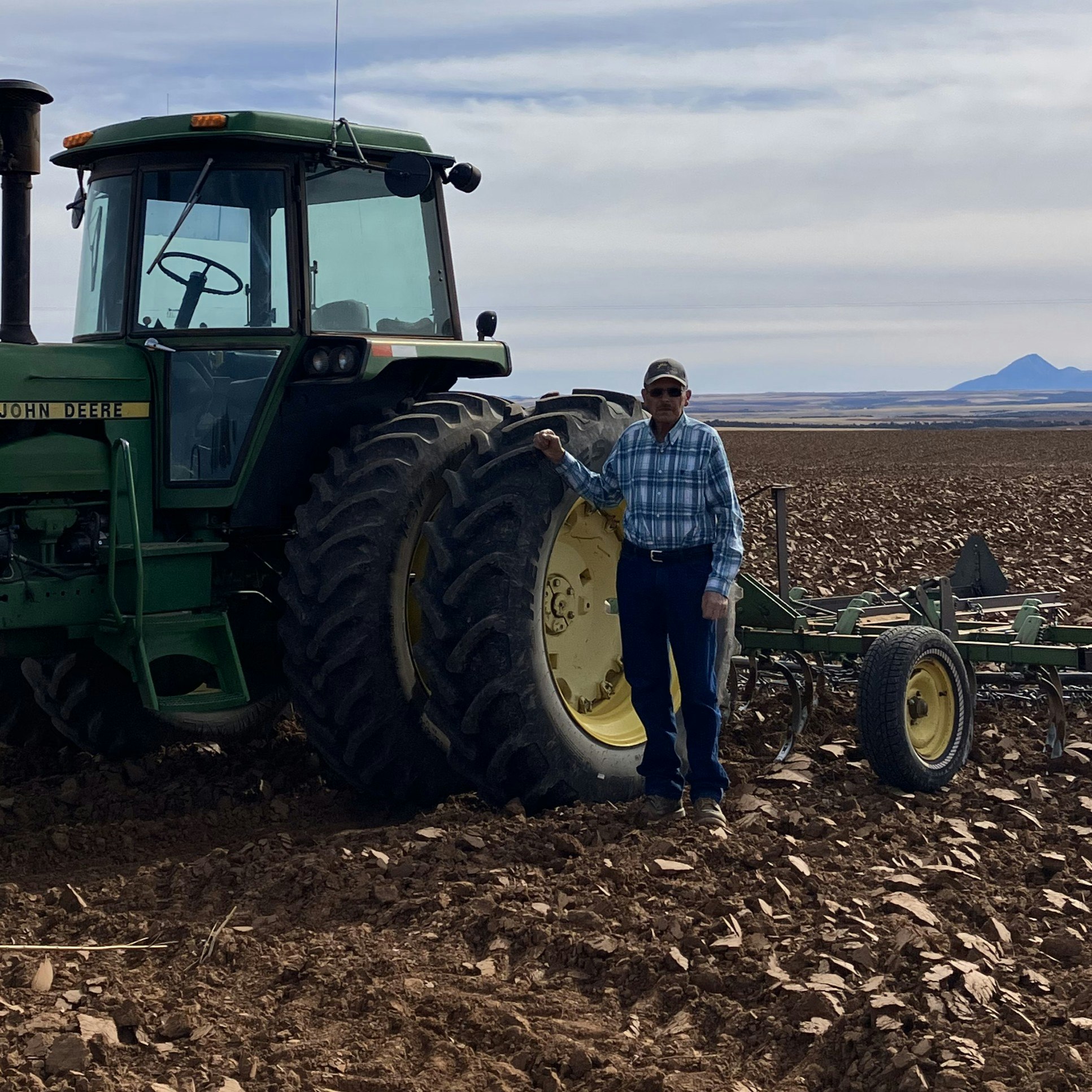 A person stands next to a John Deere tractor in a plowed field under a cloudy sky.