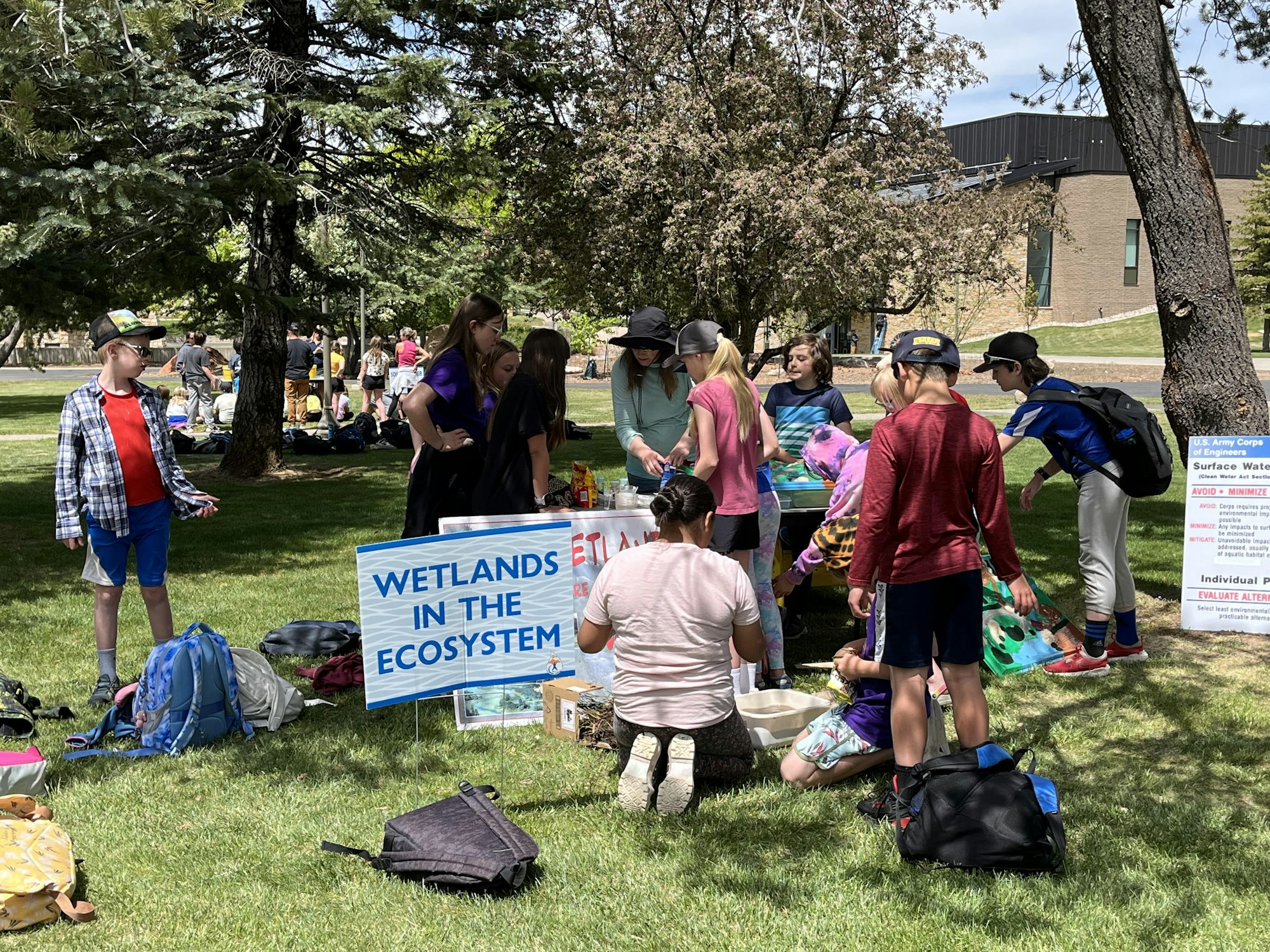 Outdoor educational event with children and adults near "Wetlands in the Ecosystem" sign, learning materials, and backpacks on grass.