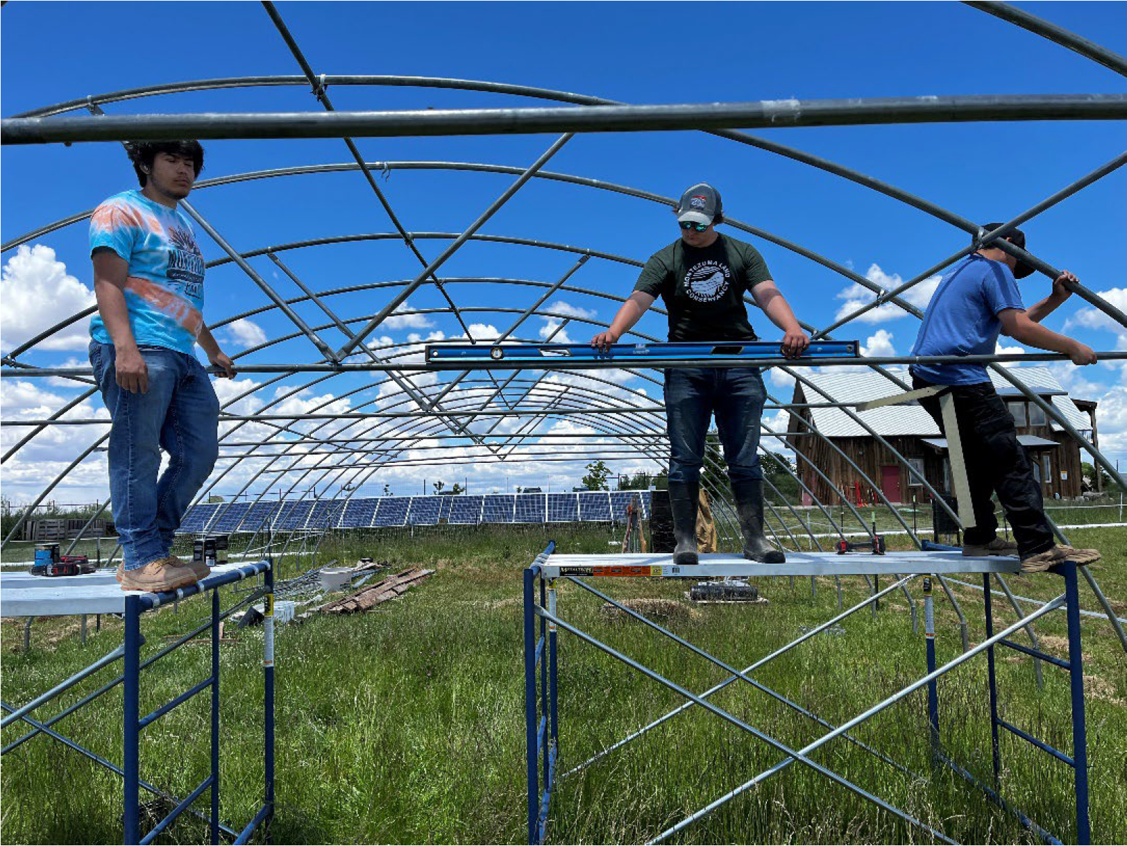Three people working on a metal structure for solar panel installation, with solar panels and a building in the background.