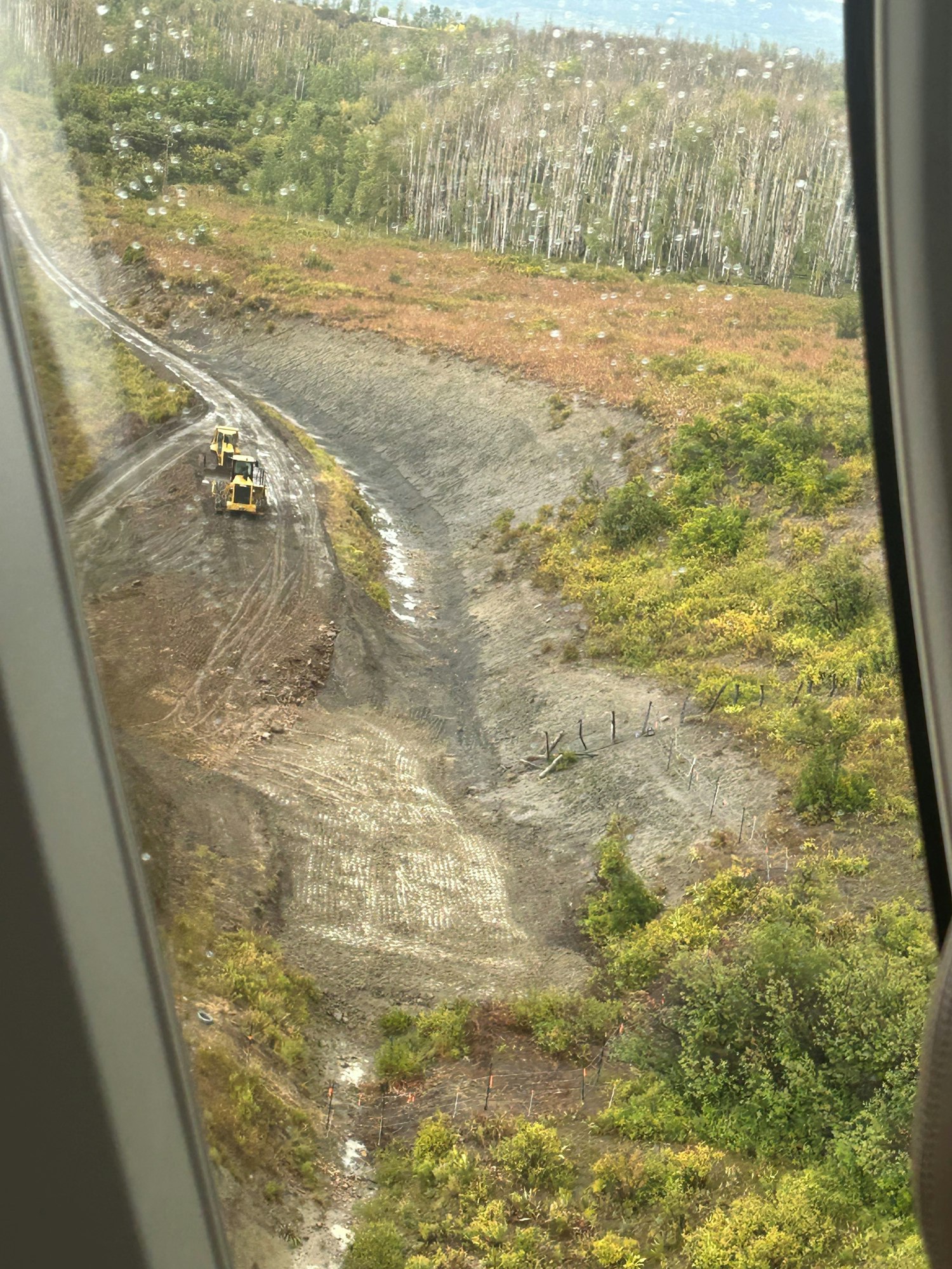 Aerial view of a deforested area with heavy machinery and wet paths, seen through a window with raindrops.