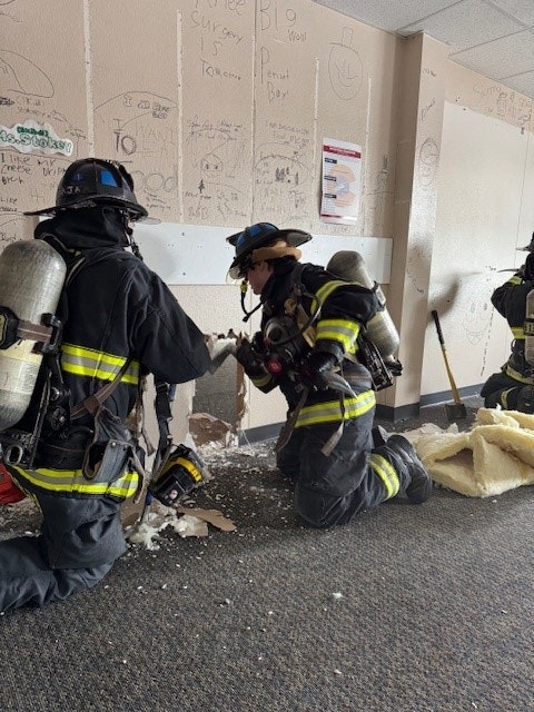 Firefighters in gear kneeling on a carpeted floor, breaking through a wall.