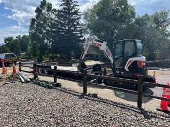 A construction site with a small excavator on the road, surrounded by traffic cones and a wooden fence. Trees and a vehicle are in the background.