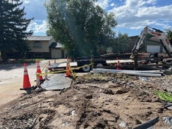 Construction site with cones, caution tape, dirt, and a small excavator near houses under a partly cloudy sky.