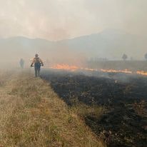 A firefighter walks near a wildfire with charred ground in the foreground and smoke against a mountainous backdrop.
