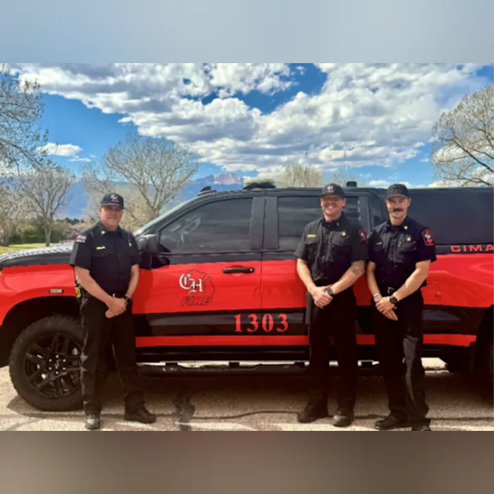 Three firefighters stand beside a red fire vehicle with a mountain backdrop and a cloudy sky.