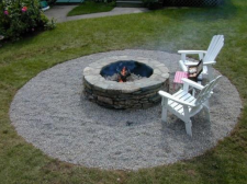 A fire pit with burning fire, two Adirondack chairs, surrounded by a gravel circle in a grassy yard.