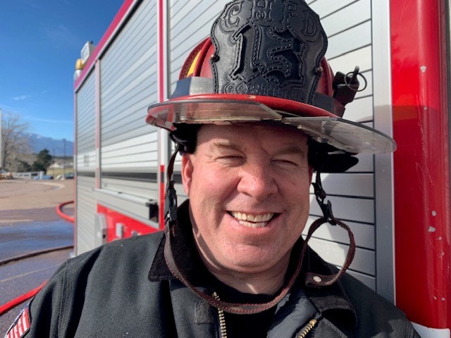 A smiling firefighter in uniform and helmet stands beside a fire truck.
