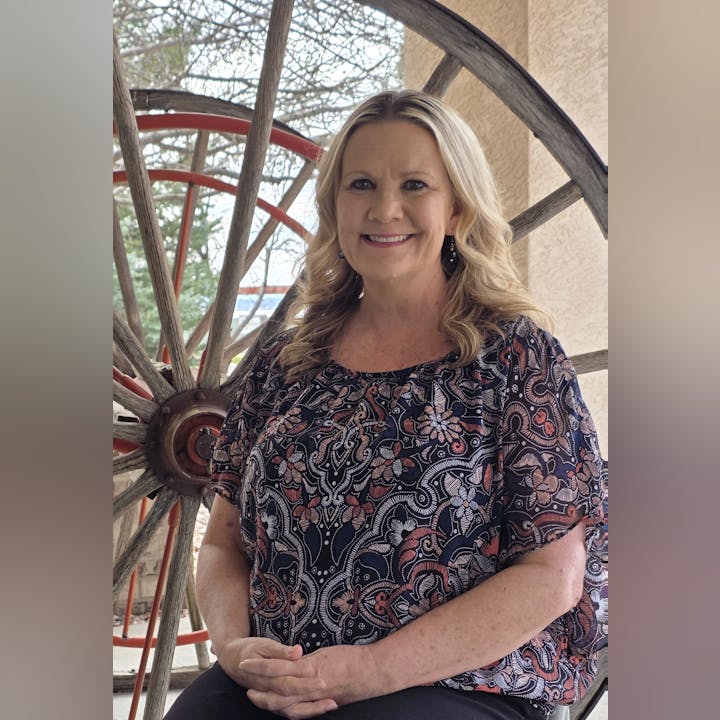 A woman smiles while sitting in front of a large wooden wheel, wearing a patterned top, with trees visible in the background.