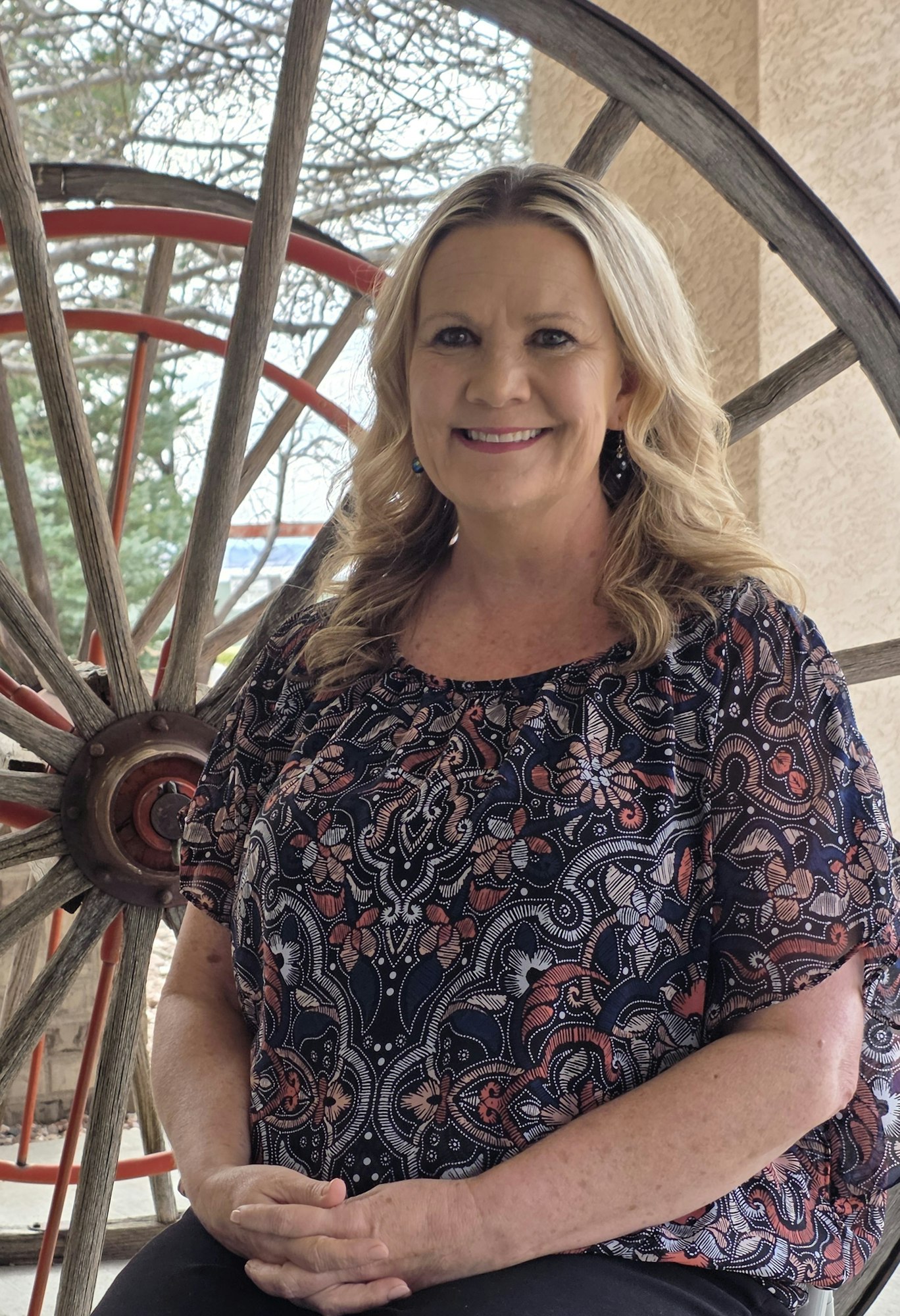A woman smiles while sitting in front of a large wooden wheel, wearing a patterned top, with trees visible in the background.