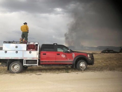 A firefighter in yellow gear stands on a fire truck, observing smoke from a fire in the distance against a cloudy sky.