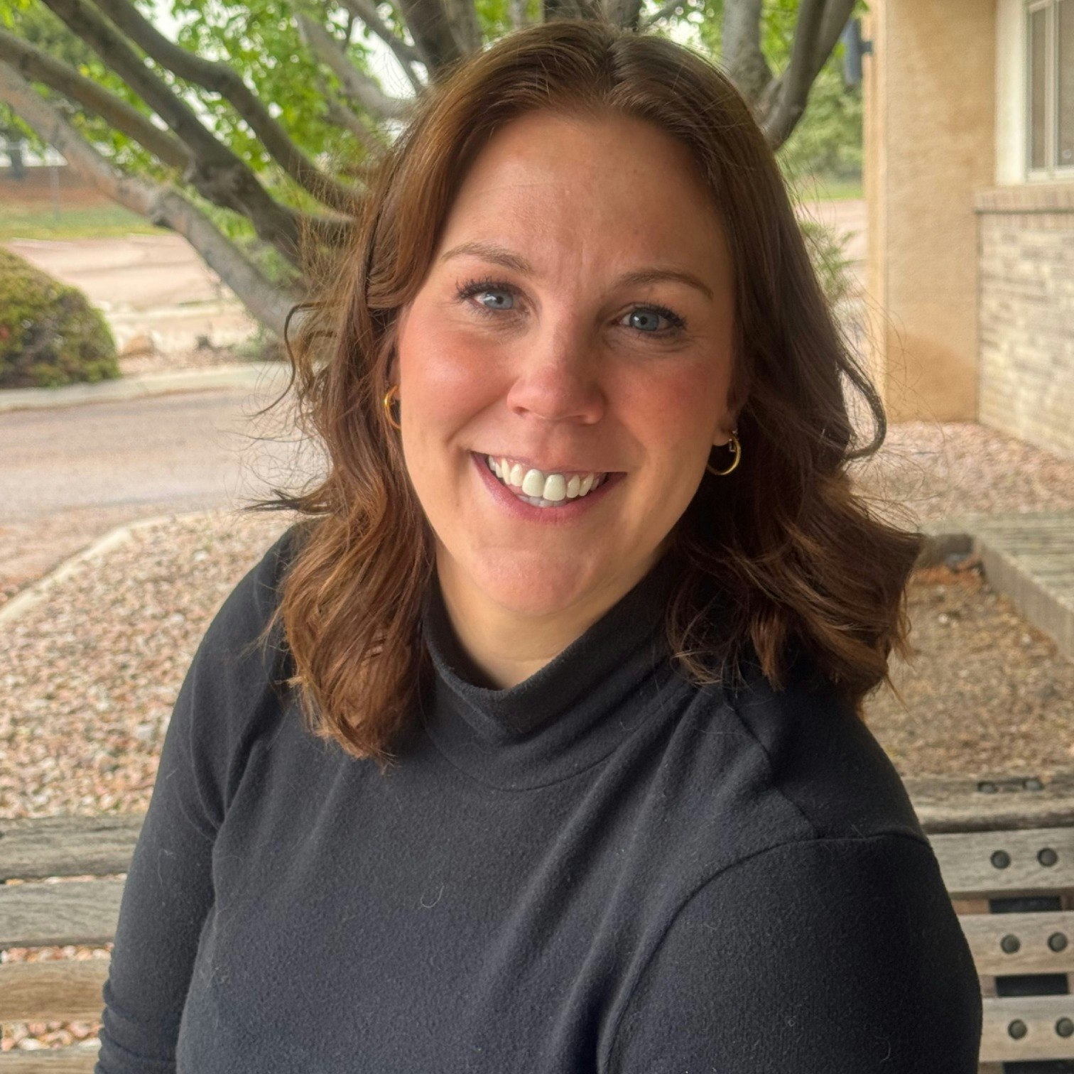 A person smiling, sitting on a bench outdoors with greenery and a building in the background.