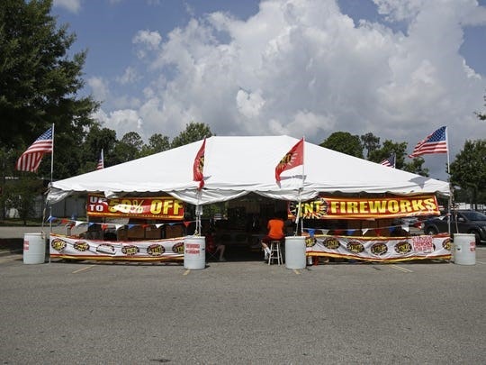 A fireworks sale stand under a white tent with banners and American flags, set up in a parking lot.