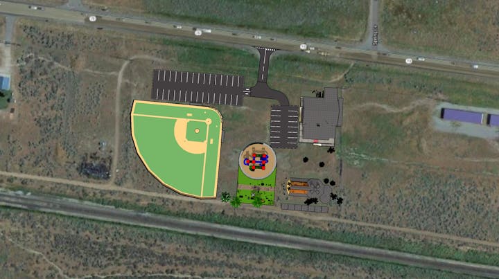 Aerial view with a baseball field, parking lots, a building, playground, and various landscape elements near a highway.
