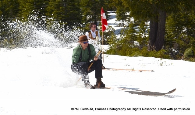 Two people are skiing on a snowy landscape, one wearing a hat while the other holds a flag in the background.
