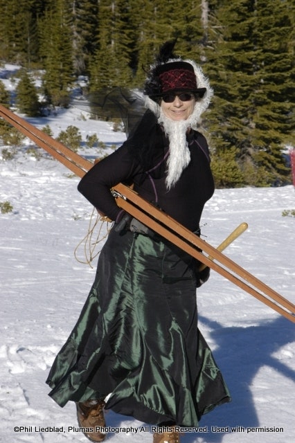A woman in a long green skirt and black top poses with skis in a snowy landscape, wearing sunglasses and a hat.
