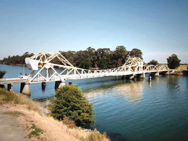 A white, truss bridge spans a calm river, surrounded by trees and greenery.