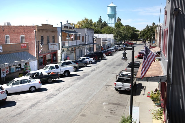 Small town street with parked cars, storefronts, and a water tower in the background.