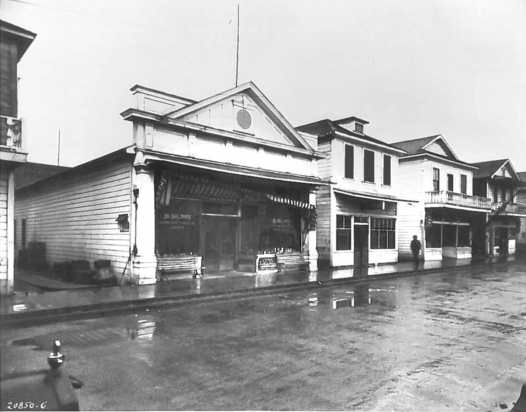A street view of old-fashioned wooden storefronts with awnings and a wet road reflecting the architecture.
