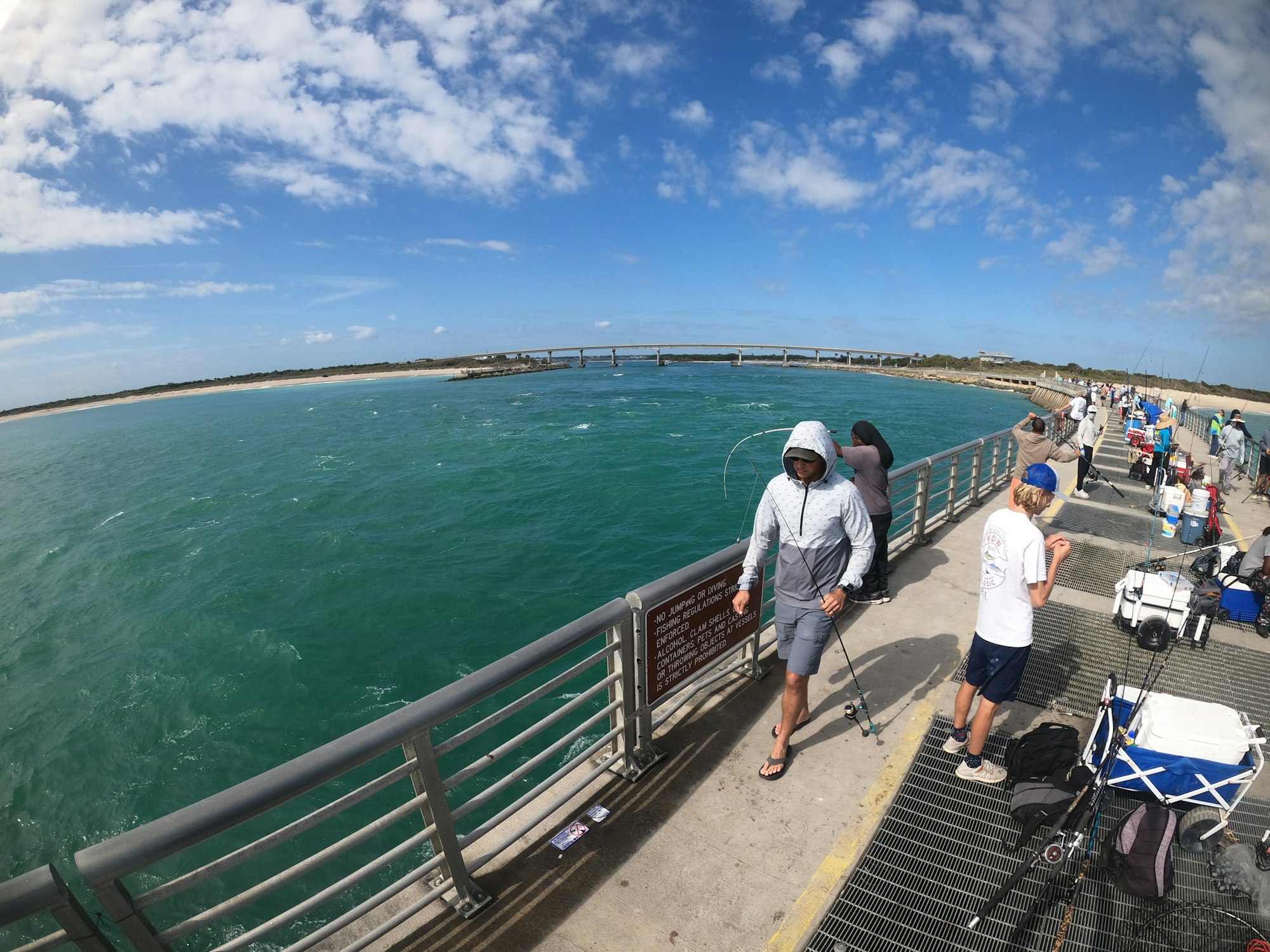 People fishing on a sunny pier with a bridge in the background.