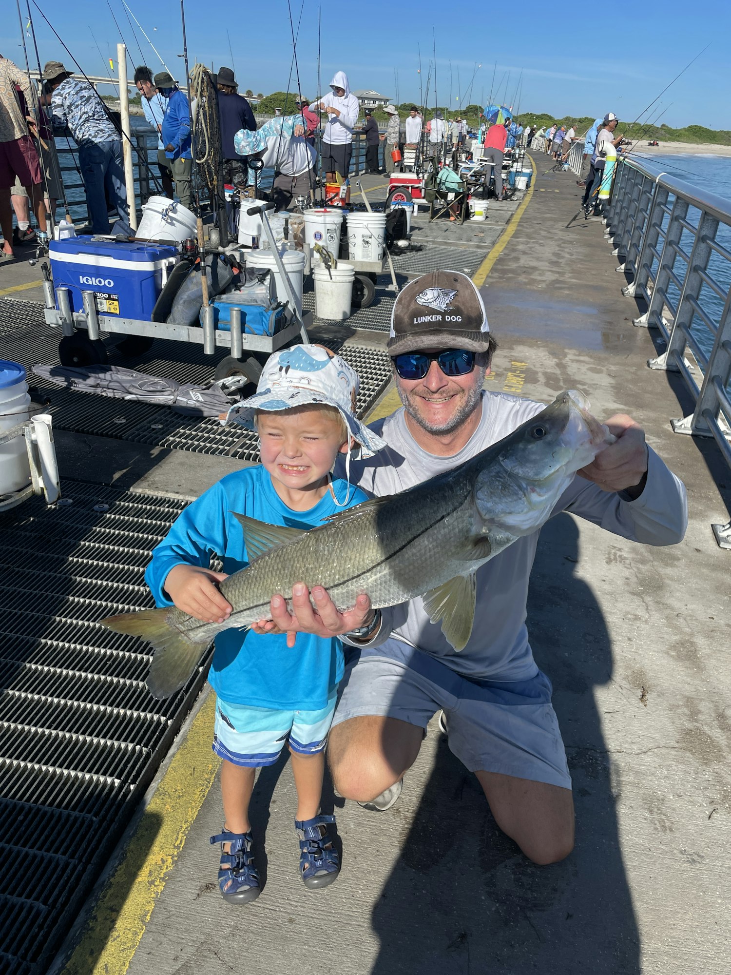 Two people holding a fish on a pier, with fishing rods and ocean in the background.