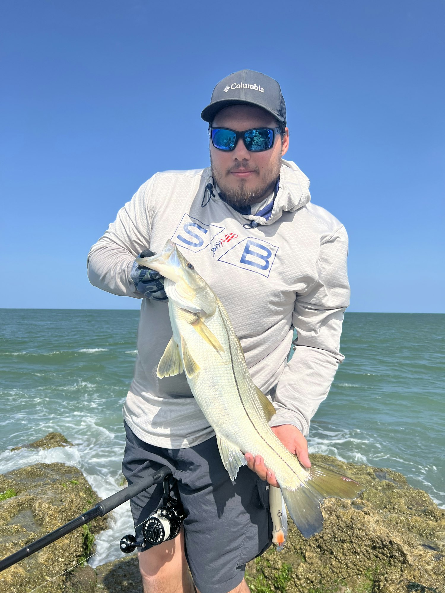 A person in sunglasses and a cap holds a fish near the ocean, on rocky terrain.