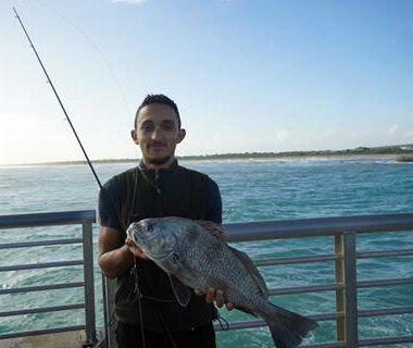 man holding fish with railing, fishing pole and ocean in background