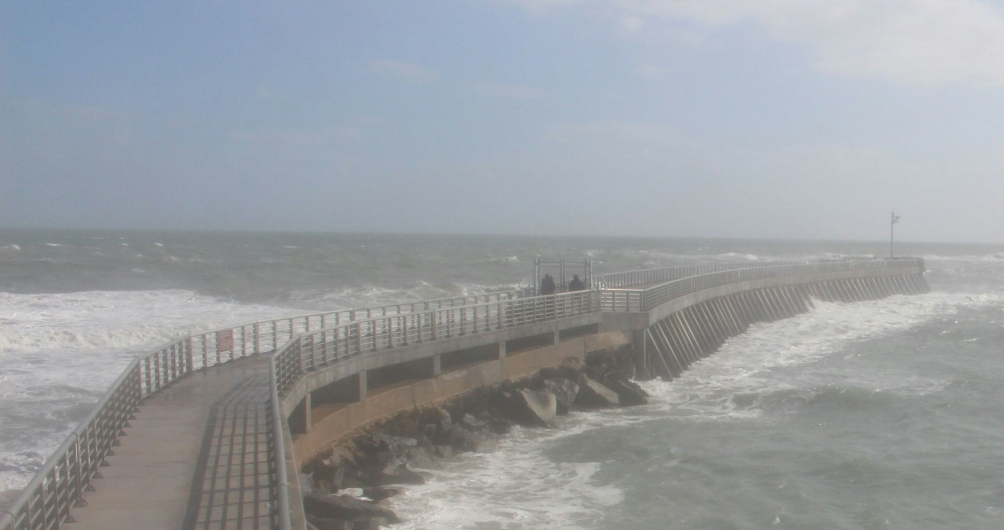 Curved pier extending into rough sea with people on it, under a hazy sky.