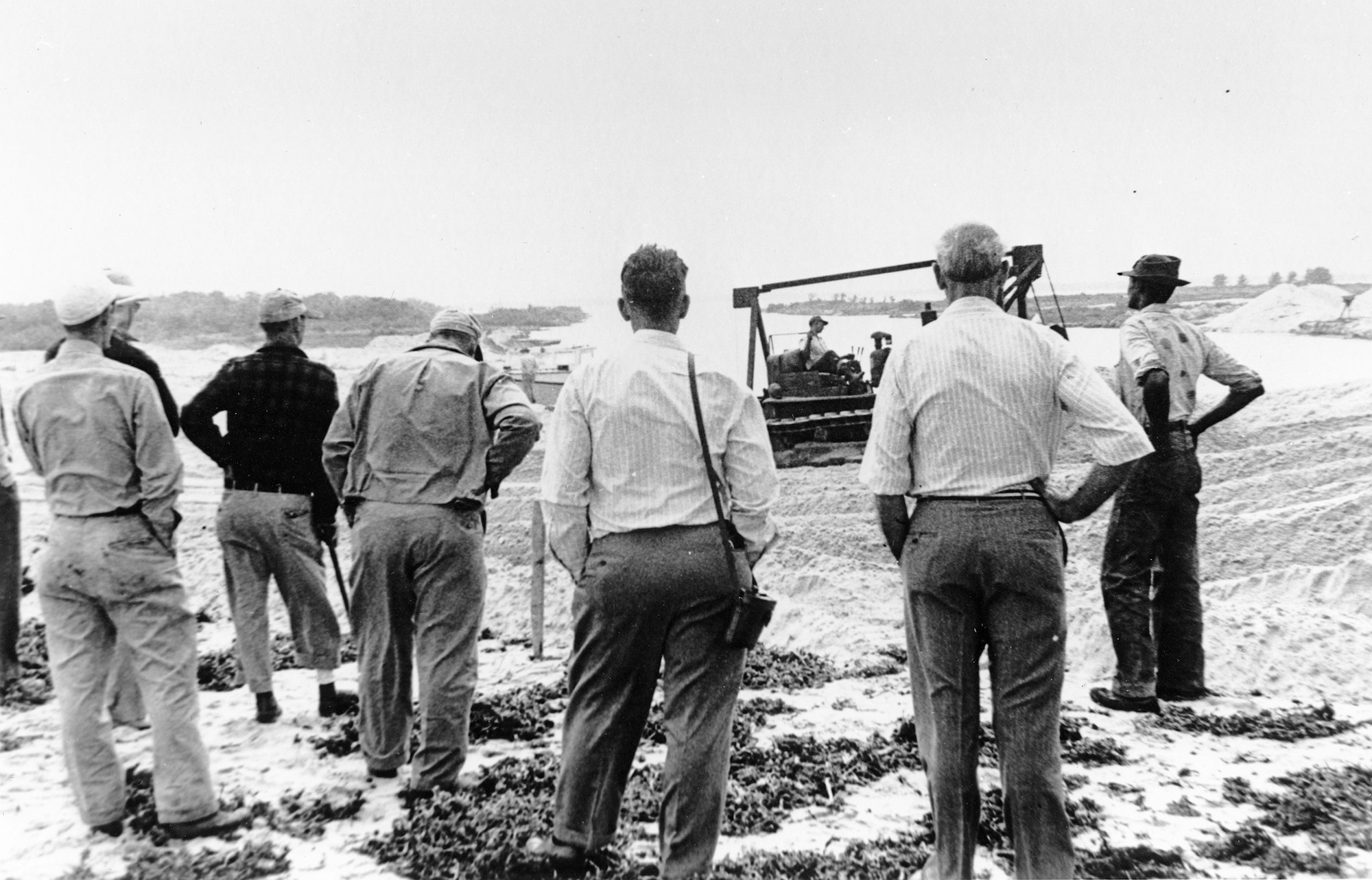 A group of men observing construction or excavation work, likely in a rural setting, with machinery in the background.