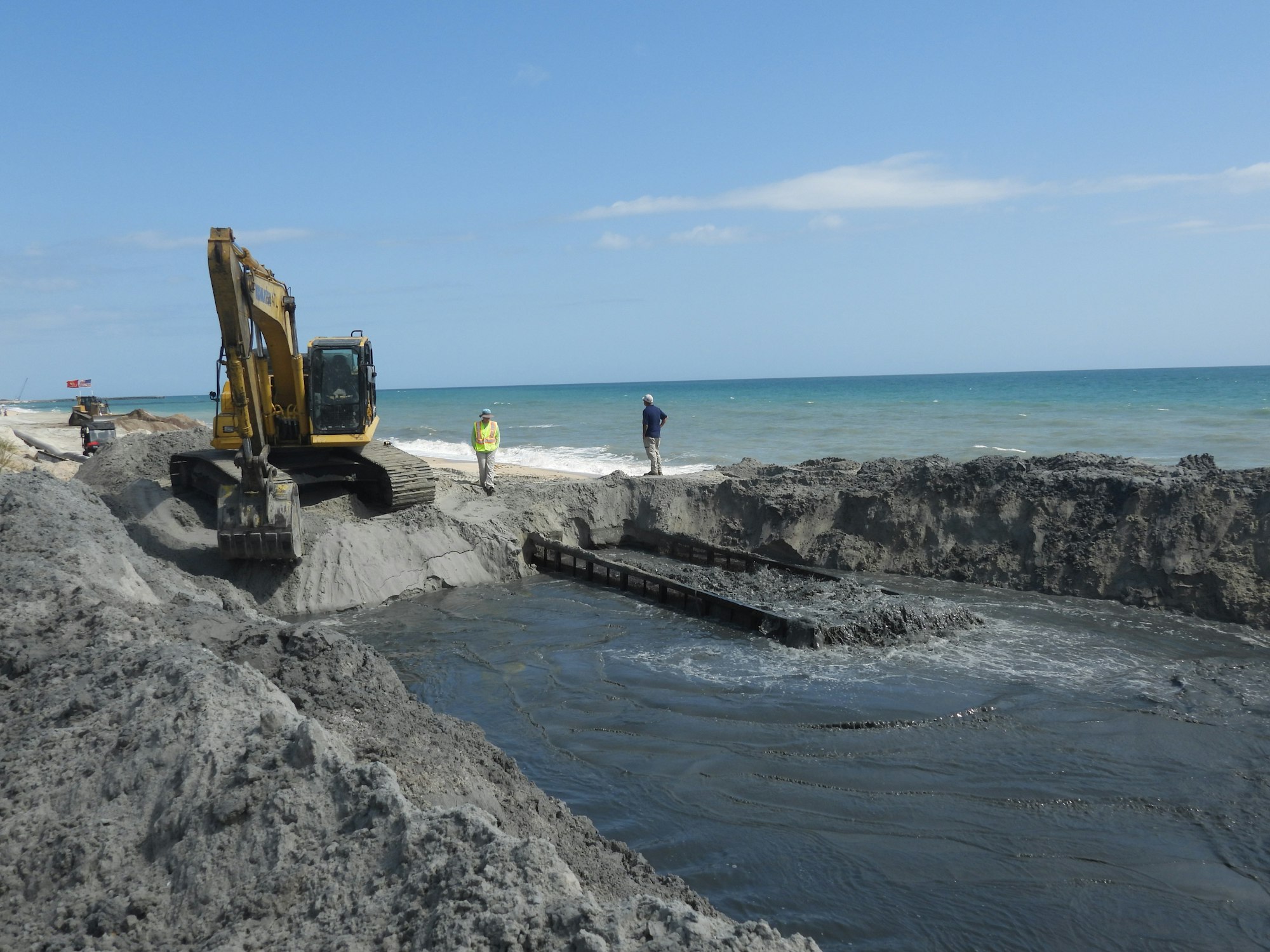 Construction work at a beach, with an excavator and workers near the shoreline, surrounded by sand and water.