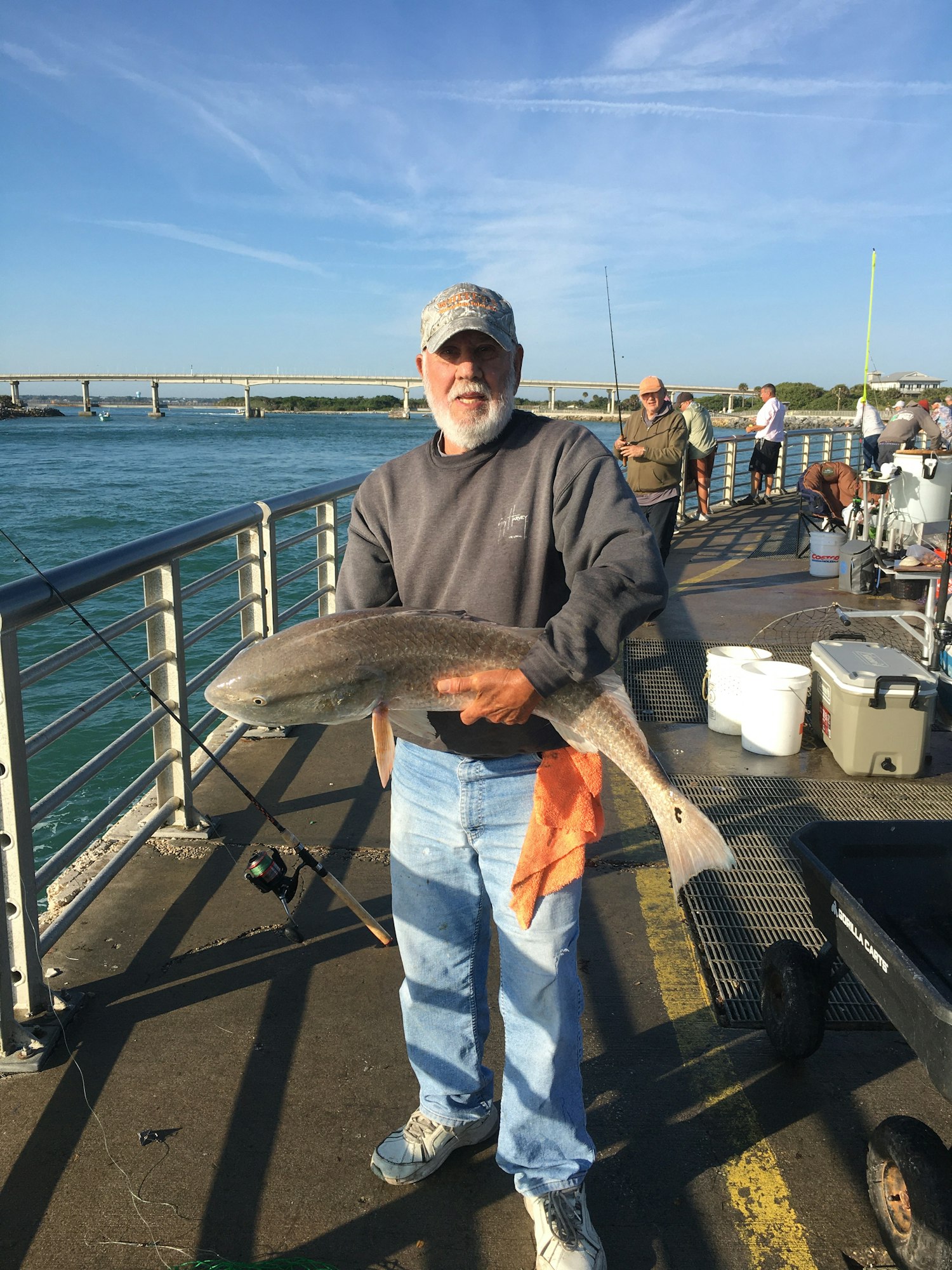 A man holding a large fish on a pier, with other fishers and a scenic water view in the background.