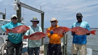 Four people on a dock showcasing large red fish, wearing casual fishing attire, with water and sky in the background.
