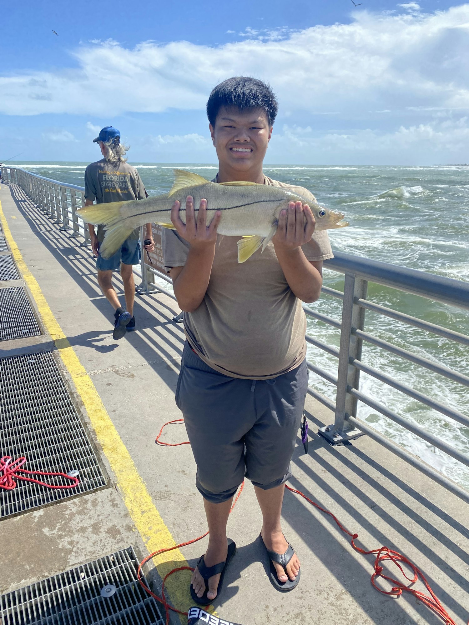 Person holding a large fish on a pier, ocean waves in the background.
